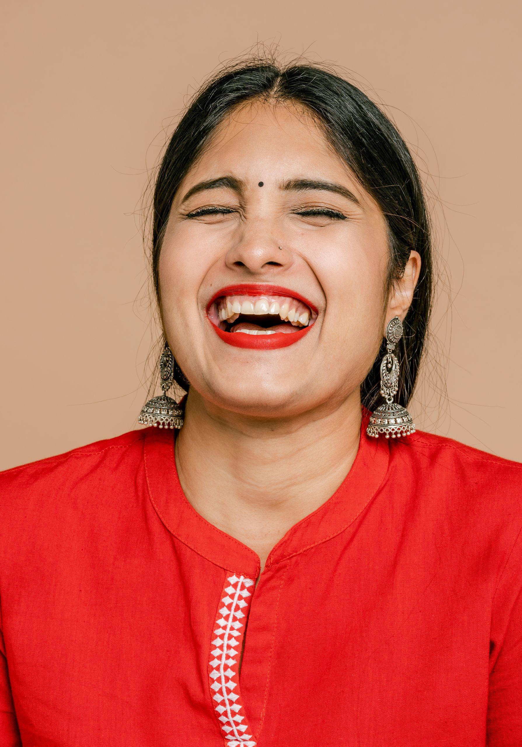 A cheerful South Asian woman laughing with red lips and traditional earrings, set against a beige background.